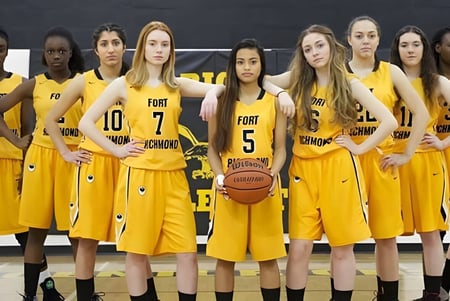 Un grupo de jóvenes mujeres en camisetas de baloncesto amarillas está juntas, una de ellas sostiene un balón de baloncesto, capturado en el campus del Fort Richmond Collegiate.