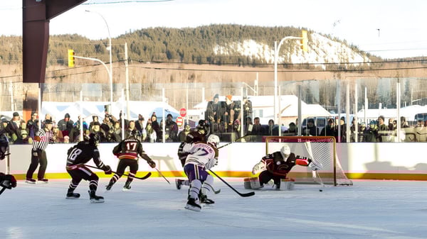 Estudiantes juegan un partido de hockey sobre hielo en el campo exterior de la Fort St. James Secondary con espectadores y montañas cubiertas de nieve de fondo.