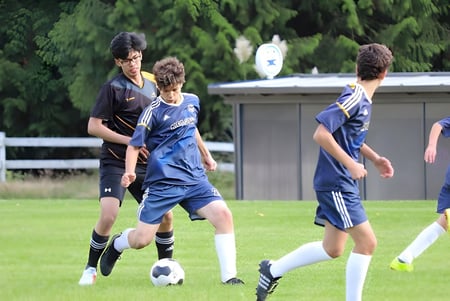 Estudiantes de la Frances Kelsey Secondary School juegan al fútbol en un campo cubierto de hierba con un edificio y árboles al fondo.