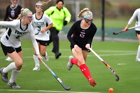 Estudiantes de The Frederick Gunn School juegan un partido de hockey sobre césped en el césped artificial con espectadores y entrenadores al fondo.