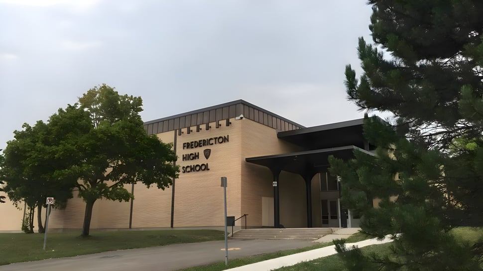 El edificio de ladrillo de la Fredericton High School con un letrero en la entrada y un camino pavimentado rodeado de árboles.