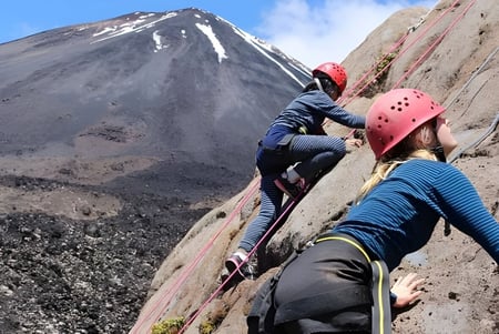 Dos estudiantes de la Freyberg High School escalan con equipo una pared rocosa con una montaña nevada de fondo.