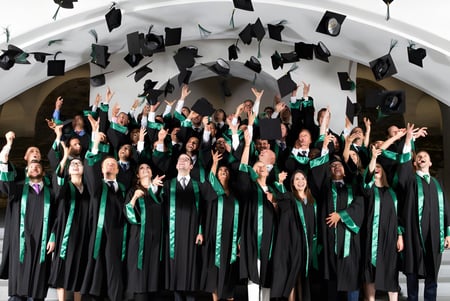 Un gran grupo de graduados de la Gallen Community School lanza sus birretes al aire frente a un arco blanco.