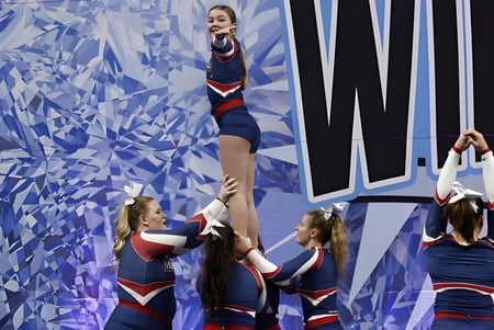 Estudiantes de la Gananoque Secondary School realizan un acrobático stunt de cheerleading frente a un fondo abstracto azul.