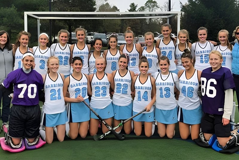 Un grupo de estudiantes de la Garrison Forest School posan en uniforme deportivo sobre un campo de césped frente a árboles y edificios.