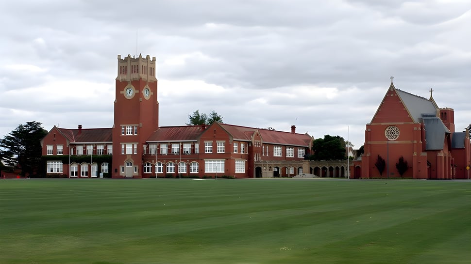 El edificio de ladrillo rojo con reloj torre de la Geelong Grammar School se encuentra en un prado verde bajo un cielo nublado.