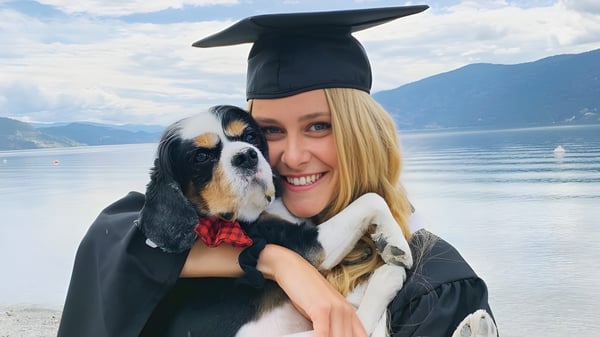 Una graduada sonriente de la George Elliot Secondary School sostiene un perro en la playa con montañas de fondo.