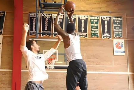 Dos estudiantes del George Harvey Collegiate Institute juegan baloncesto en un gimnasio con pancartas deportivas al fondo.