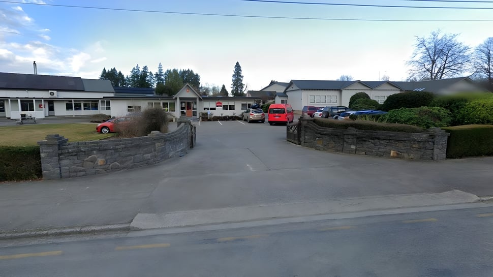 Una calle con edificios históricos y coches estacionados junto a la Geraldine High School frente a un fondo de pinos y cielo azul.