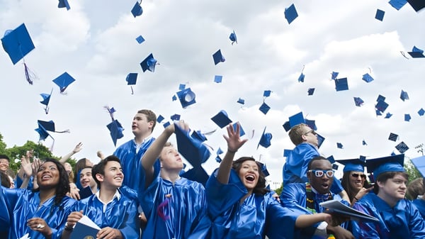 Un grupo de graduados de Gilbert Public Schools lanza su gorra de graduación azul al aire frente a un cielo nublado.