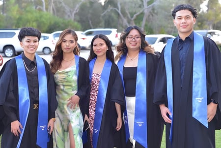 Un grupo de cinco graduados está al aire libre en el campus del Gilmore College con sus togas de graduación.
