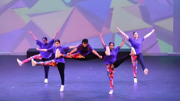 Un grupo de estudiantes de la Girrawheen Senior High School baila en el escenario frente a un fondo de formas geométricas violetas y azules.
