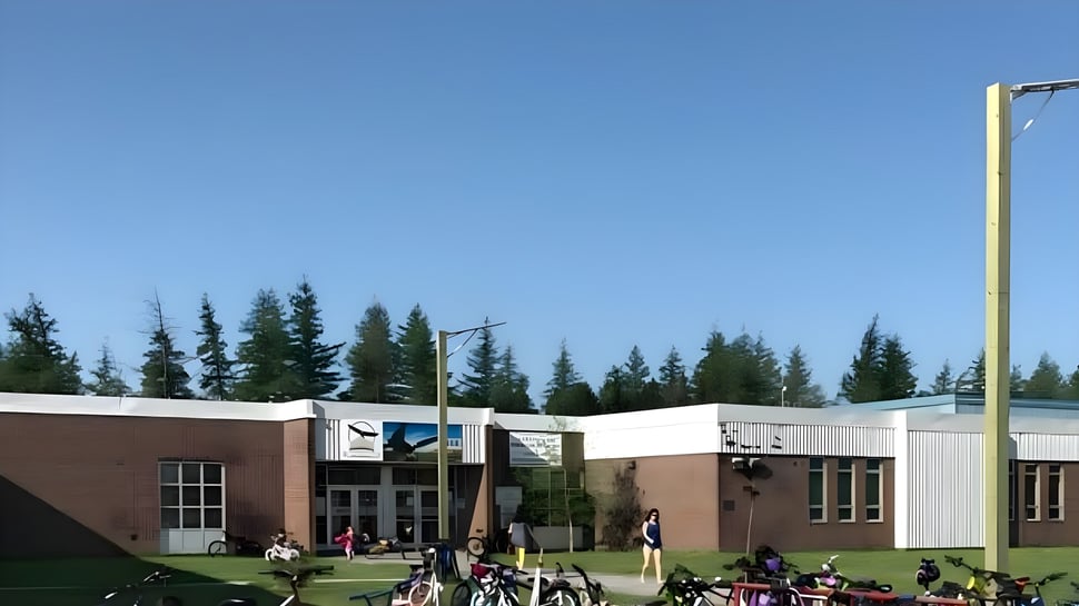 El moderno edificio escolar de la Glovertown Academy con grupos de estudiantes en el campo frente a él y un bosque al fondo.