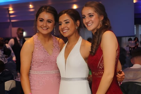 Tres alumnas de la Grange Post Primary School posan juntas en un entorno festivo con otras personas al fondo.