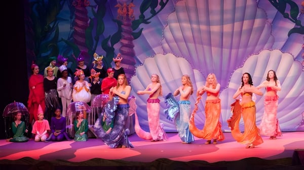 Estudiantes de Greensboro Day School están en coloridos disfraces en un escenario frente a un fondo colorido.