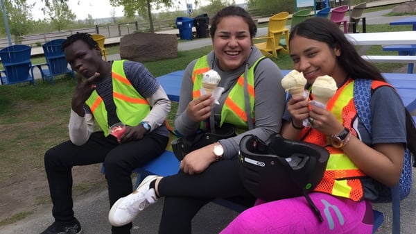 Tres estudiantes de la Greenwood Secondary School están sentados en el patio de recreo comiendo helado frente a un colorido parque infantil.