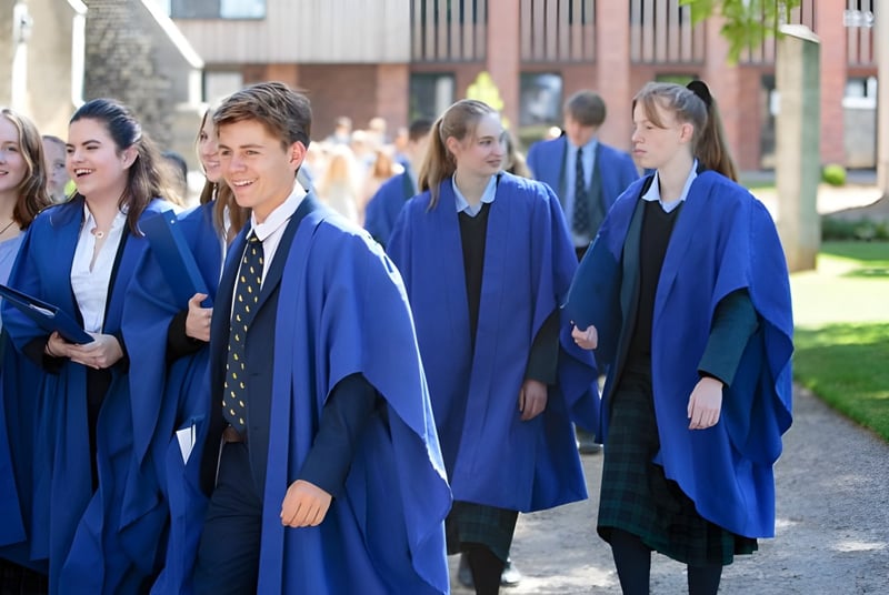 Un grupo de estudiantes de Gresham’s School con togas de graduación azules camina por un camino entre edificios y áreas verdes.