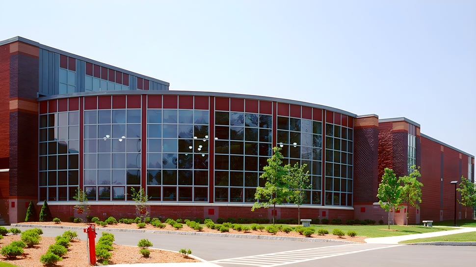 El moderno edificio de varios pisos de la Groton-Dunstable Regional High School con fachada de vidrio y ladrillo rojo está rodeado de árboles y plantas.