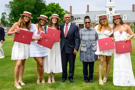 Un grupo de jóvenes mujeres en vestidos blancos y sombreros está junto a un hombre en traje en el césped de la Groton School.
