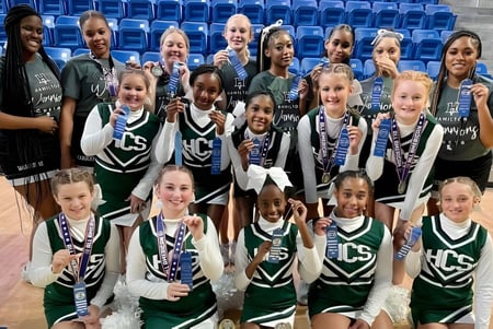 Un grupo de jóvenes jugadoras de baloncesto posan en la cancha de la Hamilton Christian Academy con gradas azules de fondo.