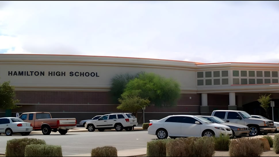 El gran edificio de ladrillo de Hamilton High School con el letrero en el edificio y un estacionamiento en primer plano.