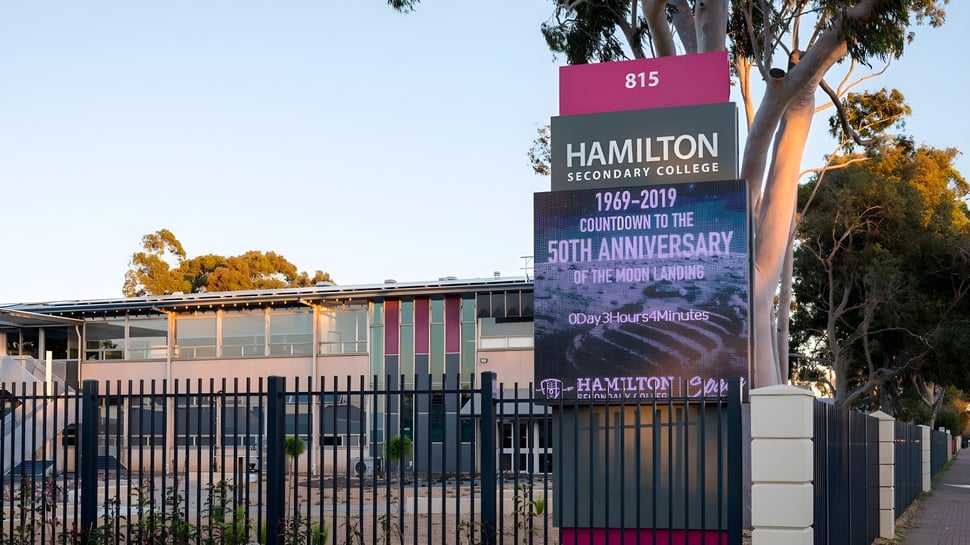 La entrada del Hamilton Secondary College con un gran cartel para la celebración de los 50 años y palmeras al fondo.