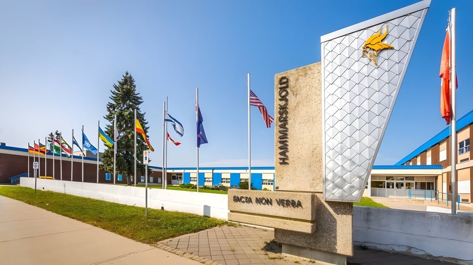 El moderno edificio escolar de varios pisos con fachada decorativa de la Hammarskjold High School está rodeado de banderas y un camino pavimentado.