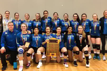 Estudiantes de la Harry Ainlay High School sostienen juntas un trofeo de voleibol en el suelo del gimnasio.