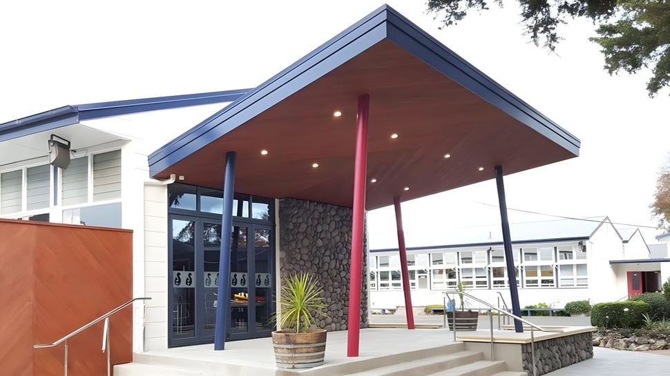 El moderno edificio de dos pisos con un toldo rojo y azul en el campus de Hastings Girls' High School está rodeado de árboles.