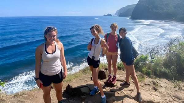 Alumnos de la Hawaii Preparatory Academy están en un acantilado mirando hacia el océano azul y una costa montañosa.