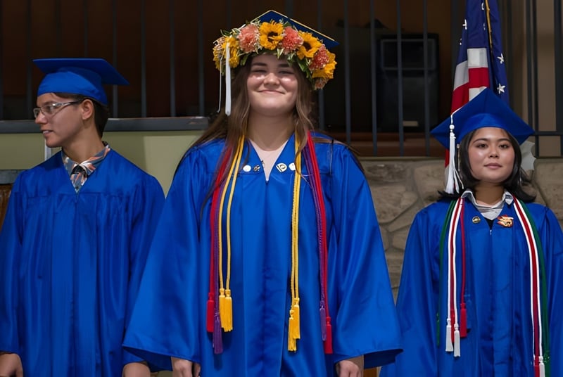 Tres estudiantes de la Hawaiian Mission Academy en togas de graduación azules frente a un edificio de piedra.