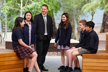 Un grupo de estudiantes y un hombre en traje están en la terraza de madera de la Heathfield High School rodeados de vegetación verde.