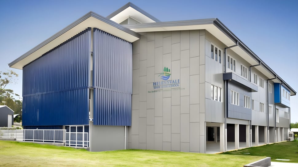 El moderno edificio de varios pisos de la Helensvale State High School con paneles exteriores grises y azules y un área de césped en primer plano.