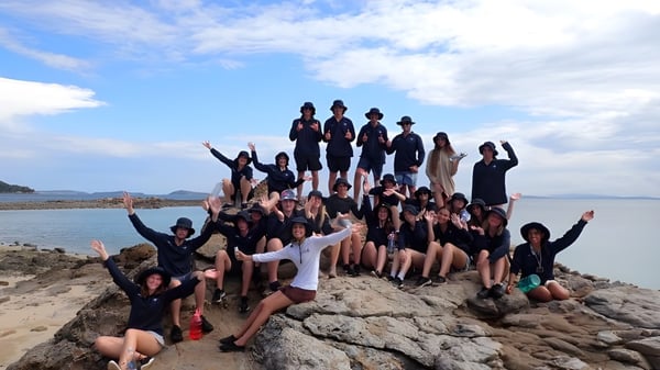 Un grupo de estudiantes de la Helensvale State High School posando en una playa rocosa con montañas y cielo azul de fondo.
