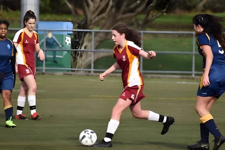 Las estudiantes del Heretaunga College juegan un partido de fútbol en el campo de césped con edificios de fondo.
