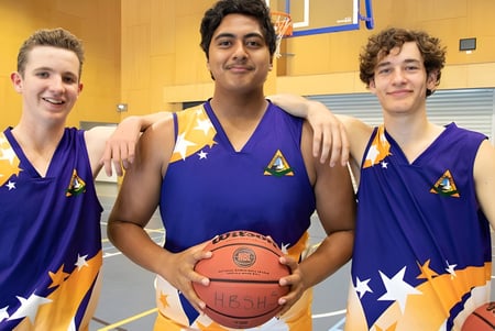 Tres estudiantes de la Hervey Bay State High School están en uniforme de baloncesto en un gimnasio y sostienen un balón de baloncesto.