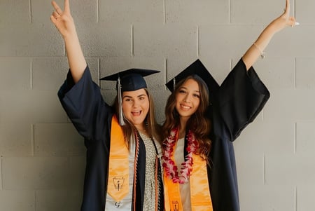 Dos estudiantes de la Higley High School celebran su graduación con los brazos en alto frente a una pared neutral.