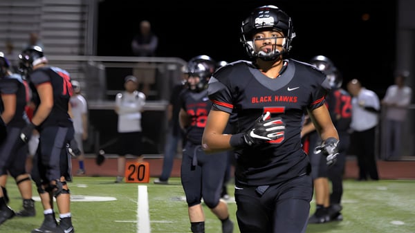 Un jugador de fútbol americano en uniforme negro y rojo está en el campo del Higley Unified School District con otros jugadores y espectadores al fondo.