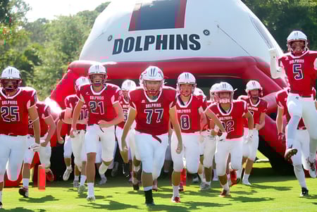 Jóvenes jugadores de fútbol americano de la Hilton Head Preparatory School están en camisetas rojas frente a un gran logo inflable de Dolphins en el campo deportivo.