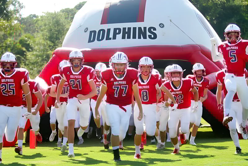 Jóvenes jugadores de fútbol americano de la Hilton Head Preparatory School están en camisetas rojas frente a un gran logo inflable de Dolphins en el campo deportivo.