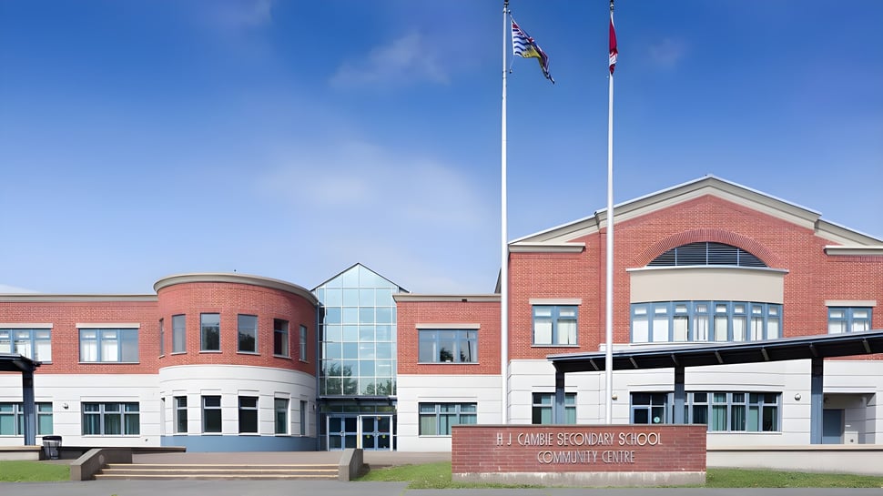 El moderno edificio de ladrillo de varios pisos de la H.J. Cambie Secondary School con entrada de vidrio y banderas frente a un cielo azul.