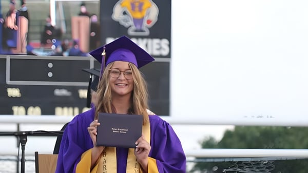 Una estudiante de la Hobart City High School lleva una toga de graduación violeta y sostiene su diploma frente a un fondo con el logo de la universidad.