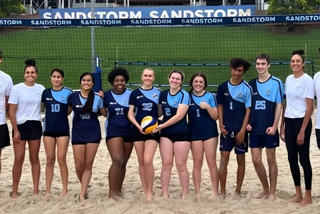 Estudiantes de la Holland Park State High School están juntos con un balón de voleibol en una playa de arena frente al cartel de Sandstorm.
