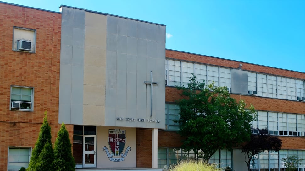 El edificio de ladrillo de varios pisos de la Holy Cross High School, con grandes ventanas y una entrada distintiva, está rodeado de árboles bajo un cielo despejado.