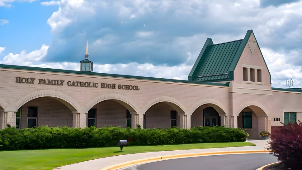 El gran edificio de ladrillo de la Holy Family Catholic High School con portal de arco y techo de torre verde está rodeado de vegetación y cielo nublado.
