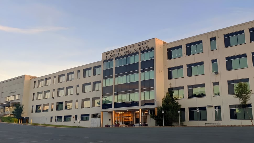 El edificio de varios pisos con ventanas de vidrio de la Holy Heart of Mary High School está rodeado de árboles y tiene un estacionamiento pavimentado en el frente.