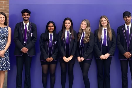 Un grupo de alumnas y alumnos de la Holy Rood High School está de pie en uniforme escolar formal frente a una pared morada con una mujer en un vestido estampado.