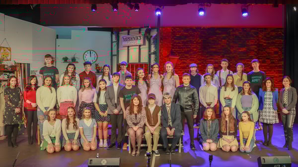 Un grupo de estudiantes está en el escenario del Holy Rosary College frente a una colorida iluminación y una pared de ladrillo.