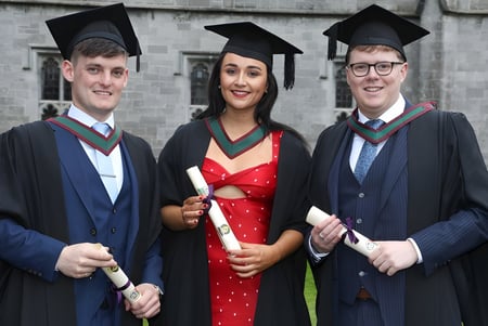 Tres estudiantes del Holy Rosary College en toga de graduación frente a un edificio de ladrillo con diplomas en la mano.