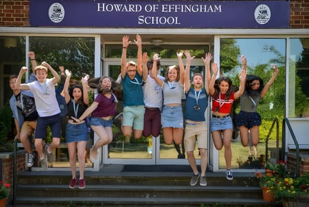 Un grupo de estudiantes está de pie con los brazos en alto frente a la Howard of Effingham School y celebra.